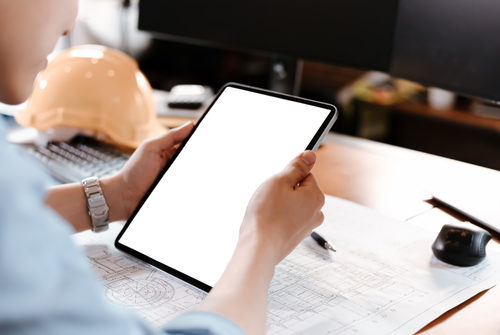 Eine Person hält ein Tablet in den Händen, im Hintergrund liegen ein Bauhelm und eine Bauzeichnung auf einem Schreibtisch