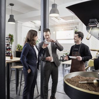Eine Frau und ein Mann in Businesskleidung stehen vor der Kaffeeröstmaschine und ein Mitarbeiter erzählt über Kaffeebohnen