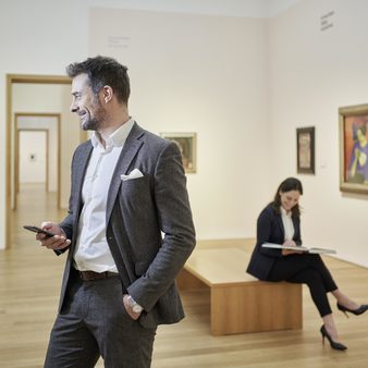 Ein Mann hält ein Handy und schaut sich ein Gemälde im Museum an, eine Frau im Hintergrund sitzt und liest ein Buch