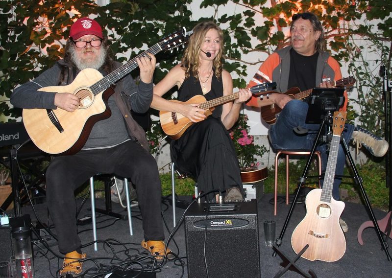 Band auf Stühlen, links kräftiger bärtiger Mann mit Gitarre, Mitte blonde Frau in schwarzem Kleid mit Ukulele und Mikro, rechts Mann mit Ukulele