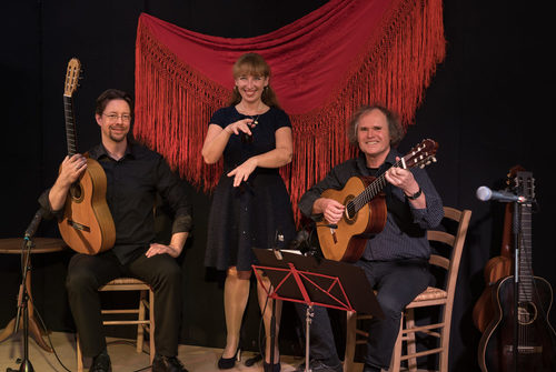 links Mann in schwarz mit Gitarre sitzend auf Stuhl, Mitte Frau in schwarzem Kleid stehend, rechts Mann in schwarz mit Gitarre sitzend