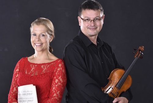 Links steht eine Frau im roten kleid mit Musiknoten in der hand und rechts ein mann mit einer Violine.