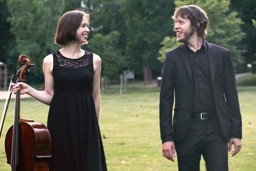 Zwei Menschen in schwarz und schick gekleidet stehen in einem Park. Links eine Frau mit braunen, kurzen Haaren und einem Cello und rechts ein Mann mit braunen Haaren. Sie schauen sich an.