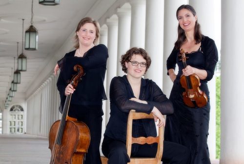 Drei Frauen in schwarz gekleidet stehen in den Wiesbadener Theaterkolonaden. Die Linke Frau hält ein Cello, die Frau in der Mitte sitzt auf einem Stuhl, welcher mit der Lehne zu uns steht und die Frau rechts hält eine Geige. Alle drei Frauen Lächeln.