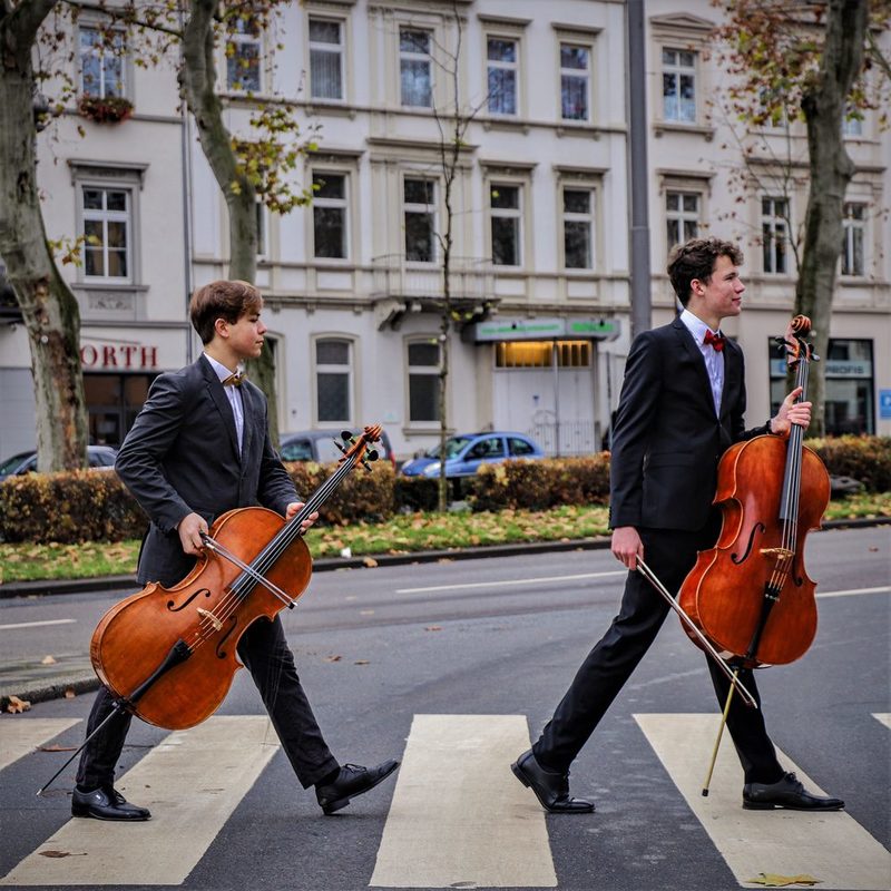 zwei junge Cellisten, die mit im Anzug mit ihrem Cello in der Hand einen Fußgängerweg überqueren