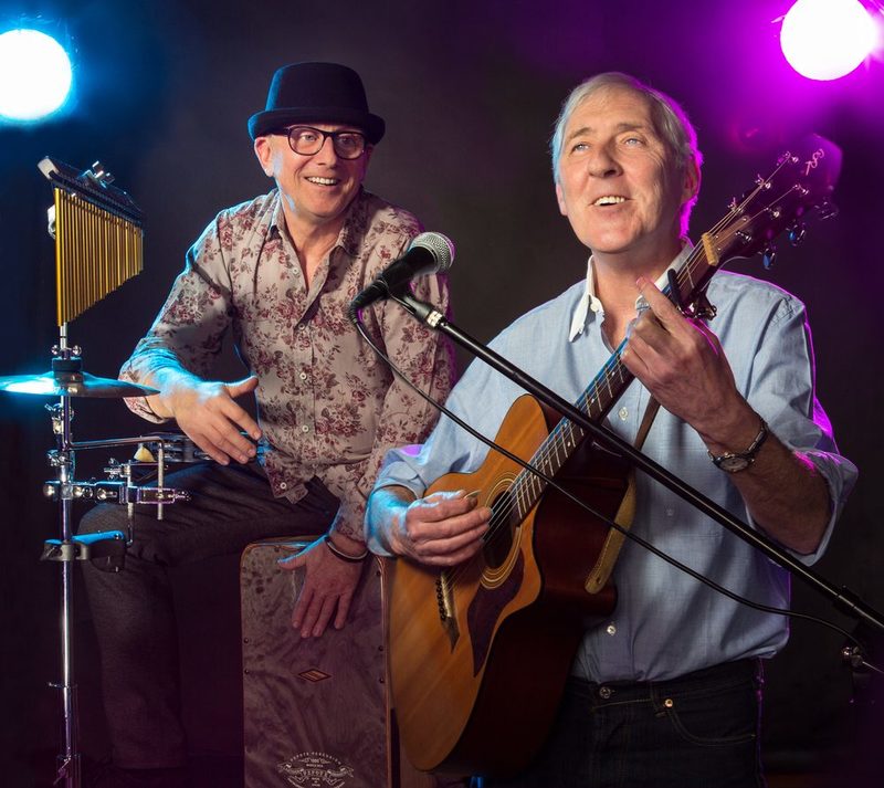 Links sitzt ein Mann mit einem Hut an den percussions und rechts sitzt ein Mann an der Gitarre. Vor ihm steht ein Mikrofon und es sieht so aus als würde er singen.